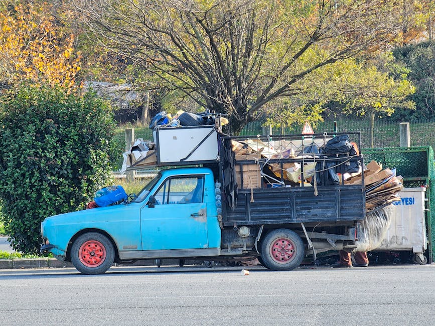 A small blue flatbed truck parked on the side of a street, fully loaded with various household items including cardboard boxes, plastic containers, and furniture wrapped in fabric or plastic covering. The load extends high above the truck's cab and is secured with ropes and straps, ready for a home relocation or furniture transport. The vehicle is positioned next to a curb with a large hedge on one side and a tree with autumn foliage in the background. The setting appears to be in daylight with natural lighting, and the truck is connected to a metal waste collection container at the rear. Occasionally used by Man with Van Woodside for removals and moving services, this scene illustrates the process of loading and transporting household belongings during a house move or furniture transport.