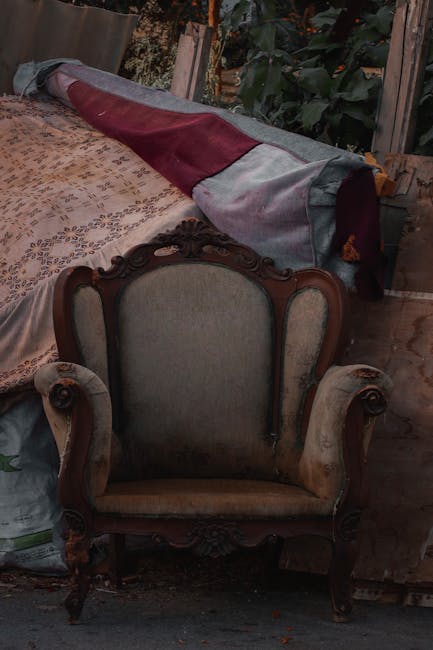 An antique upholstered armchair with wooden carved details on its frame, positioned on a wooden floor inside a property. Behind the chair, there is a large rolled-up fabric item, possibly a blanket or cover, placed on top of a table or surface covered with a patterned cloth. The setting appears to be a cluttered room used for packing or storage, with wooden furniture and visible greenery or garden outside through an open door or window. This image reflects elements involved in home relocation and packing, relevant to furniture transport and moving services offered by Man with Van Woodside. The environment is lit with natural light, emphasizing the textures of the fabric, wood, and upholstery, capturing a typical scene during furniture moving or packing for a house removal.