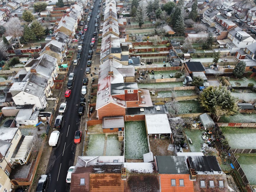 An aerial view of a residential neighbourhood showing a row of terraced houses and detached homes lined along a narrow street with parked cars on both sides. The roofs of the buildings are dusted ligh