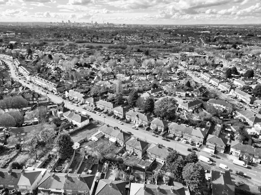 An aerial view of a residential neighbourhood showing a row of terraced houses and detached homes lined along a narrow street with parked cars on both sides. The roofs of the buildings are dusted ligh