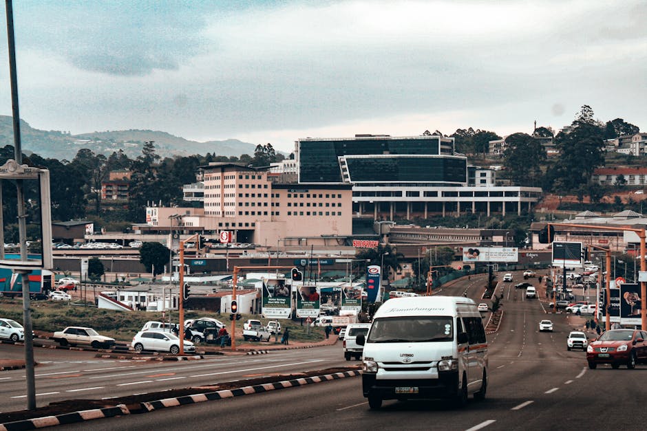 A busy urban street scene showing multiple lanes of traffic with various white and silver cars, including a prominent white van parked or moving along the road. Behind the road, there is a commercial area with signs, billboards, and storefronts. In the background, there are modern buildings with large glass windows and a mix of flat and sloped roofs, set against a backdrop of green hills and trees under a cloudy sky. The scene depicts typical logistics and transportation elements involved in house removals and furniture transport, with the van and vehicles on the road aligned with activities such as home relocation, packing and moving, and loading processes, relevant to the services provided by Man with Van Woodside.