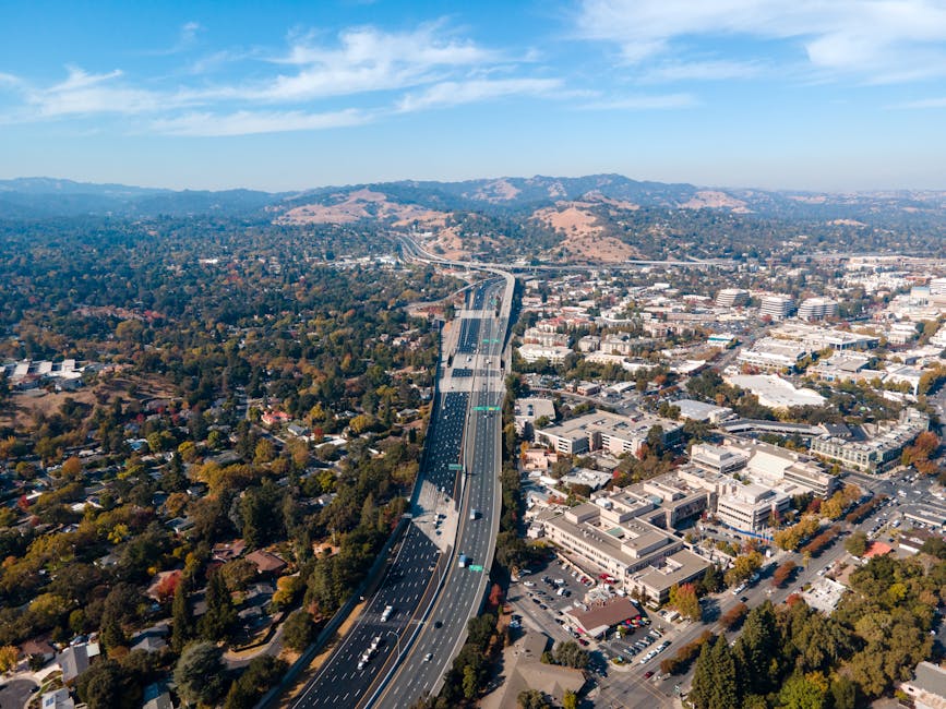 A panoramic view of a residential neighbourhood in Woodside during daylight, featuring a variety of houses with different roof styles, including tiled and flat roofs, surrounded by many green trees and shrubs. The street is visible winding uphill among the homes, with some parked cars along the curb. In the background, a mountain range with lush greenery rises under a partly cloudy sky, providing a scenic backdrop that highlights the area's natural landscape. This setting may be part of a property or street scene related to home removals or furniture transport in the context of relocating to Woodside from Addiscombe, as coordinated by Man with Van Woodside. The overall scene emphasizes the peaceful residential environment and diverse housing architecture typical of the area, suitable for a detailed description supporting moving and packing services.