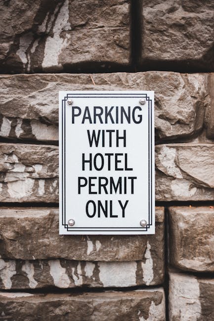 A rectangular white metal sign mounted on a textured stone wall, displaying the message 'PARKING WITH HOTEL PERMIT ONLY' in bold black uppercase letters. The sign is affixed with four screws—one in ea