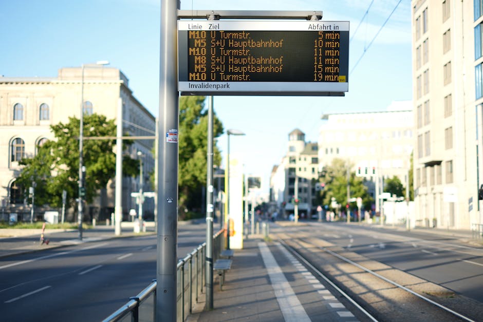 A digital tram timetable display board mounted on a metal pole on a city sidewalk, showing tram departure times to various destinations in yellow text against a black background. The display is located at a tram stop next to tram tracks that run parallel to the pavement, with a metal railing separating the waiting area from the rails. Behind the tram stop, there are multi-story buildings with a mix of modern and historic architecture, alongside trees with green foliage. The surrounding area appears quiet with minimal traffic or pedestrians during daylight hours. This scene illustrates typical urban transport infrastructure for residents preparing for travel or moving in a city such as Woodside, supporting home relocations involving transport arrangements and logistical planning, similar to those managed by Man with Van Woodside.