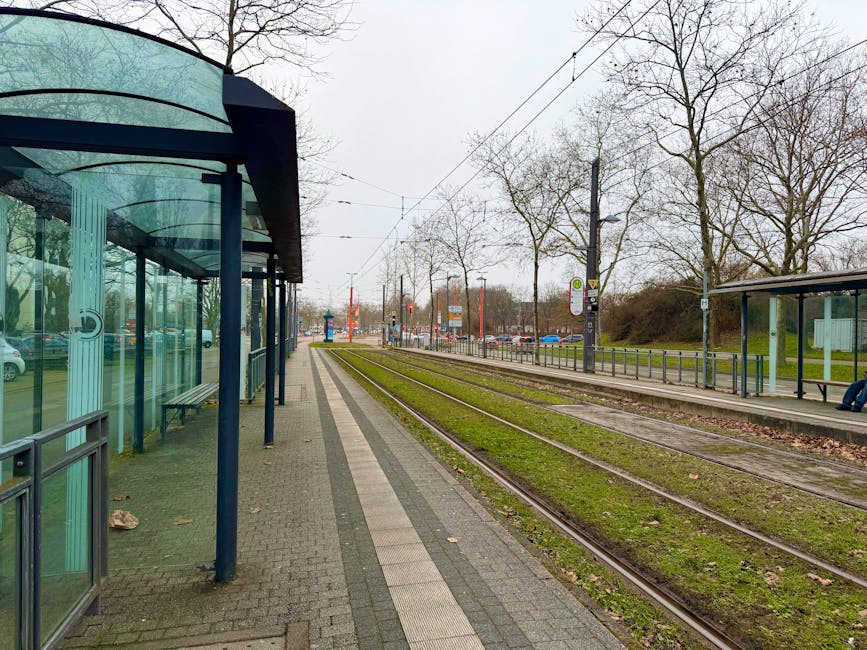 An exterior view of a tram stop adjacent to a residential street, taken on a cloudy day. The tram shelter features a curved, transparent glass roof supported by dark blue metal poles and includes a bench inside, with some cardboard and packaging materials visible on the pavement. Parallel tram tracks run along the grassy area beside the shelter, leading into the distance. Overhead electric wires are suspended above the tracks, supported by tall poles. In the background, there are leafless trees, signage, parked cars, and a traffic light. The scene appears to be part of a home relocation process, with a focus on moving logistics and transport preparations in the context of house removals near Woodside Tram Stop, as indicated by the page title.