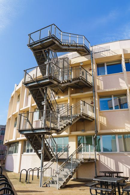 The image shows the exterior of a multi-storey residential building with a metal fire escape staircase attached to the facade, featuring multiple landings and black railings. The building has large rectangular windows, and the walls are painted light cream or beige. The staircase ascends from the ground level, which has bicycles locked to a rack, ascending to the upper floors, with a clear view of the blue sky overhead. The setting appears to be situated on pavement or a communal outdoor area often associated with house relocations and property management. Inside, the building likely contains apartments, and the staircase provides access between floors. The scene relates to property access issues during home relocations, and the image subtly supports services related to removals and furniture transport, particularly handling challenging staircase access scenarios. It demonstrates the importance of professional moving services like those offered by Man with Van Woodside, especially when navigating staircase access in flats during packing and moving processes.