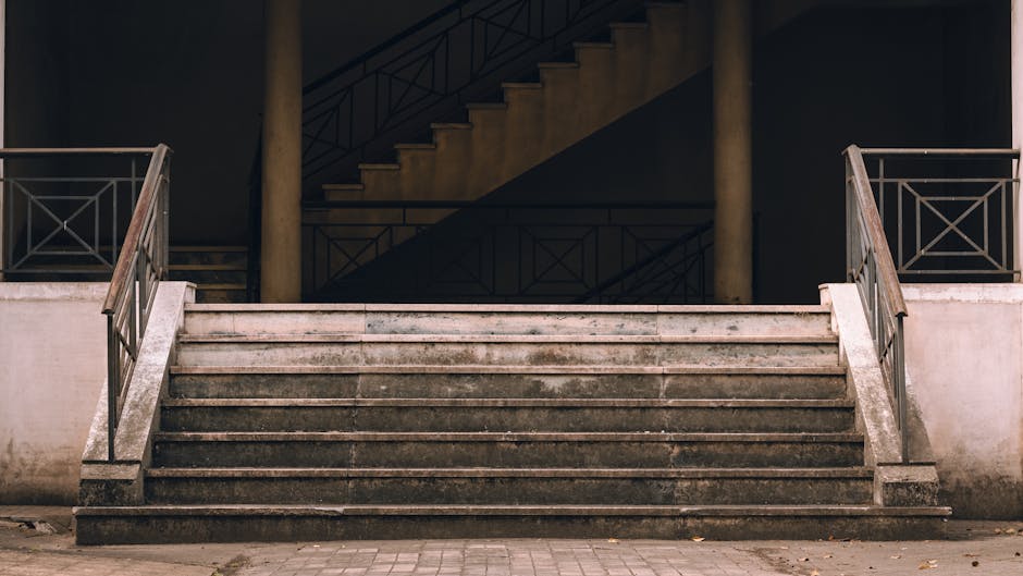 A wide view of an outdoor stone staircase leading up to the entrance of a residential building, with metal railings on each side and concrete steps that appear worn and slightly stained. The staircase is situated on a paved pavement, and the building behind features a dark interior space visible through an open doorway or window at the top. Inside, visible objects include cardboard boxes, some wrapped in plastic or fabric, indicating packing or moving preparations. A man with a van, branded as Man with Van Woodside, is engaged in a home relocation process nearby, possibly loading or unloading items. The scene reflects a move involving furniture and packed boxes, with the staircase presenting access challenges often encountered during house removals within flats, emphasizing the importance of safe loading and transportation solutions.