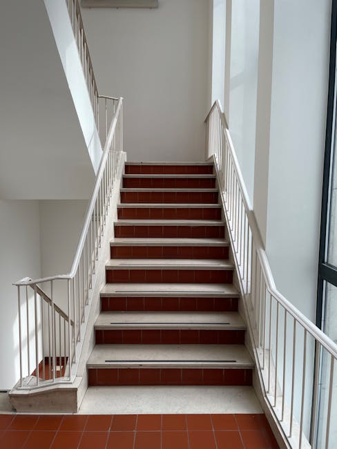 The image shows the exterior of a multi-storey residential building with a metal fire escape staircase attached to the facade, featuring multiple landings and black railings. The building has large rectangular windows, and the walls are painted light cream or beige. The staircase ascends from the ground level, which has bicycles locked to a rack, ascending to the upper floors, with a clear view of the blue sky overhead. The setting appears to be situated on pavement or a communal outdoor area often associated with house relocations and property management. Inside, the building likely contains apartments, and the staircase provides access between floors. The scene relates to property access issues during home relocations, and the image subtly supports services related to removals and furniture transport, particularly handling challenging staircase access scenarios. It demonstrates the importance of professional moving services like those offered by Man with Van Woodside, especially when navigating staircase access in flats during packing and moving processes.