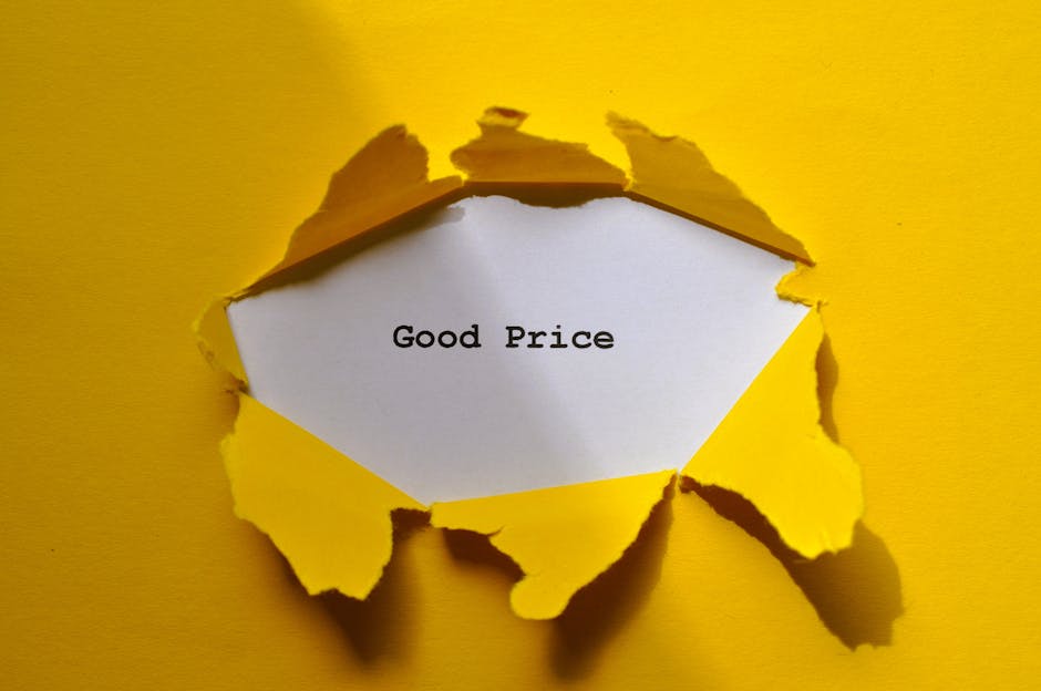 A close-up photograph of a torn piece of yellow paper revealing a white card beneath it, which displays the text 'Good Price' typed in a black monospaced font. The torn yellow paper has irregular, jagged edges and is partially peeled back, creating a circular hole through which the white card is visible. The background is a flat yellow surface that matches the torn paper, and the lighting highlights the texture and shadows created by the torn edges. This image visually represents affordability or value in the context of house removals or moving services, with the torn paper symbolising unveiling good value deals associated with companies like Man with Van Woodside, which provides removals and relocation services. The scene emphasizes the process of packing, unpacking, or transparent pricing during home relocation or furniture transport.