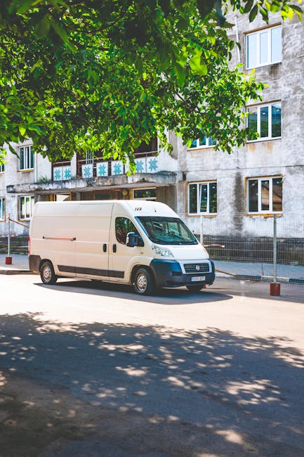 A white commercial van with a high roof is parked on the street outside a residential building, which has multiple windows and a weathered concrete facade. The van is positioned close to the pavement, with its side door closed and no visible markings or branding. Overhead, a large leafy green tree partially shades the vehicle, casting shadows on the road. In the background, a metal fence runs along the edge of the pavement, and the building's entrance is visible beneath a small overhang. This scene captures the typical environment for house removals or furniture transport, suggesting the vehicle is prepared for a home relocation or moving service. Man with Van Woodside, a professional removals company, might be involved in coordinating this logistics operation, including loading furniture and boxes into the van in preparation for transport. The overall setting highlights a residential area suitable for same-day removals in Woodside, with ample space for loading and unloading items for a furniture move or packing and moving process, indicating efficient planning for home relocation services.