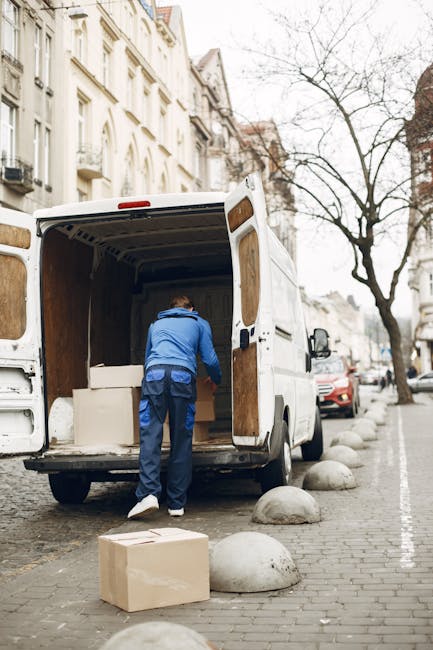 A white Iveco moving van is parked on a narrow urban street with tall, multicoloured residential buildings on either side. The back doors of the van are open, revealing the interior filled with cardboard boxes, wrapped furniture, and packing materials such as plastic wrap and blankets, indicating a home relocation process. Some boxes are stacked inside near the entrance of the vehicle, while others are placed on the pavement nearby. The street pavement is flanked by potted plants, street bollards, and a few parked bicycles. Leafless trees with thin branches cast shadows across the scene, and the late afternoon light creates warm tones on the buildings. The image captures the commercial and logistical aspects of furniture transport and packing during a same-day removals service, as provided by Man with Van Woodside, in preparation for an efficient moving and loading process.