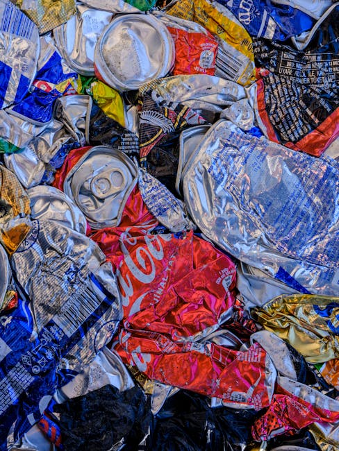 A close-up image showing a pile of crushed aluminium beverage cans, predominantly in silver, blue, red, and yellow, with some displaying visible brand labels, including a partially visible Coca-Cola can. The cans are tightly compacted together, with some overlapping each other, and their surfaces reflecting light. The background includes a few crumpled plastic packaging materials and possibly shredded paper or cardboard as part of waste or recycling materials. This scene is set outdoors on a pavement or ground surface. The presence of the cans and packaging suggests a focus on waste disposal or recycling processes, relevant to house removals or moving services offered by Man with Van Woodside, especially in contexts involving clearing or disposing of household waste before or after a move.