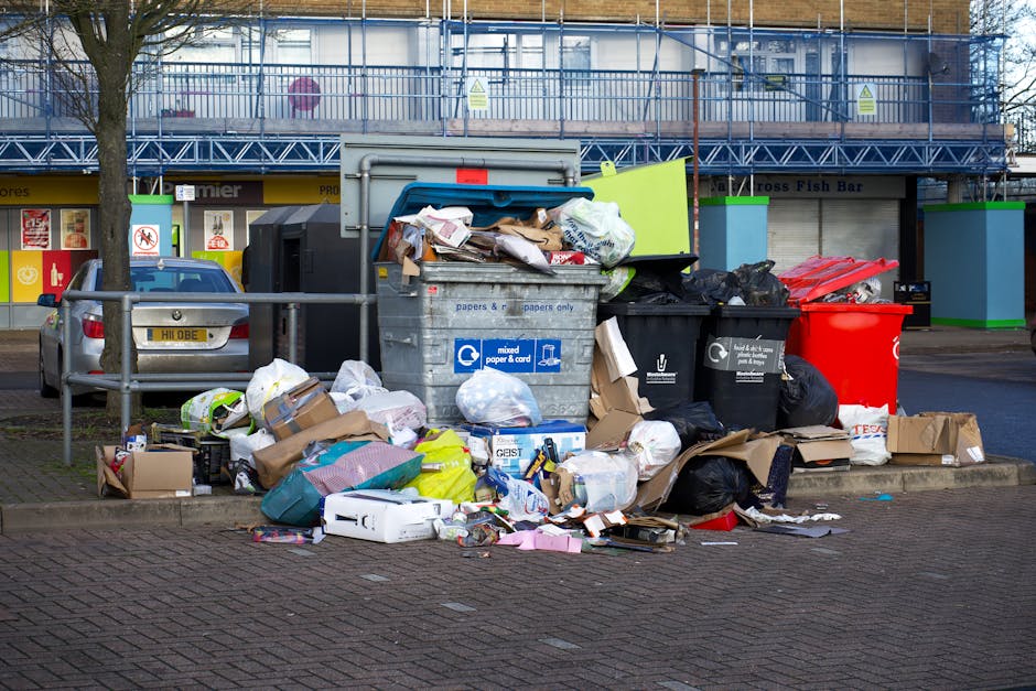 A collection of overflowing waste and recycling bins placed on a paved sidewalk in front of a building with large windows and a scaffolding structure. The waste containers include a mixed paper and cardboard bin, a black general waste bin, a black recycling bin, and a red bin, all filled with various rubbish such as paper, cardboard boxes, plastic bags, and packaging materials. Additional waste and packaging are scattered around the bins on the ground, including flattened boxes, plastic wraps, and paper. Nearby, there is a parked silver car partially visible, with a fence separating the parking area from the bins. This scene reflects improper waste disposal in an urban setting, which relevant for furniture transport and home relocation contexts, as managed by services like Man with Van Woodside.