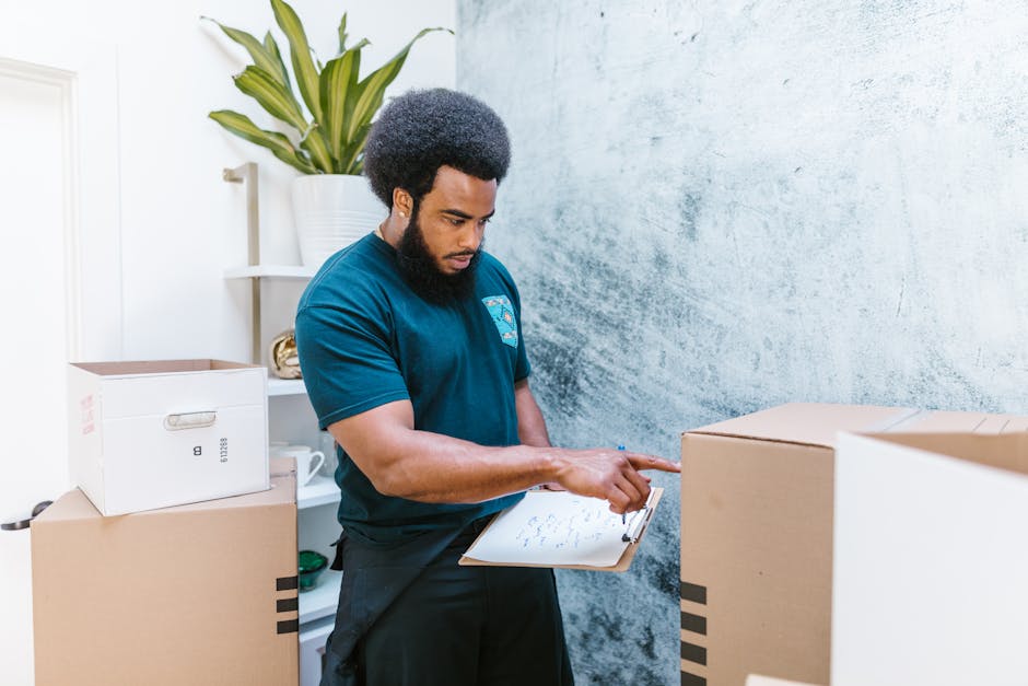 A man with dark skin and an afro hairstyle, wearing a dark blue T-shirt, is inside a room surrounded by moving boxes made of cardboard and plastic wrap. He is holding a clipboard and appears to be inspecting or organizing the items, possibly making notes related to packing or the home relocation process. Behind him, there is a white shelf with a potted plant and some books or objects. The room has a textured light grey wall and is well-lit, reflecting a professional environment for house removals. The boxes are stacked on the floor and are likely packed for transportation by [COMPANY_NAME], which specializes in furniture transport and packing services as part of their house removal operations. This image illustrates the planning and documentation stage of a professional moving service.