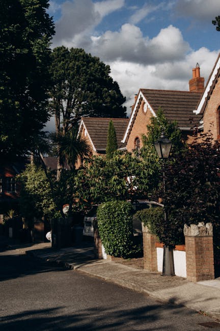 A residential street scene showing a row of brick houses with pitched roofs, some with chimneys, surrounded by tall trees and well-maintained garden foliage. In the foreground, a pavement runs parallel to the properties, with a brick wall and a pathway leading to the entrances. Partially visible in the lower left corner, a vehicle is parked on the street, and a street lamp can be seen mounted on a pole near one of the houses. The sky above is partly cloudy with patches of blue, suggesting daytime. This scene exemplifies a typical suburban environment that might be involved in home relocation or furniture transport activities coordinated by Man with Van Woodside during a house moving process.